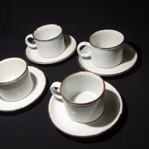 May include: Set of four white ceramic coffee cups with brown rims and matching saucers. The cups and saucers have a speckled glaze.