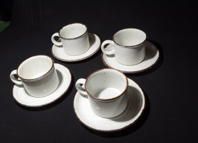 May include: Set of four white ceramic coffee cups with brown rims and matching saucers. The cups and saucers have a speckled glaze.