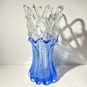 Puede incluir: Un jarrón de vidrio azul con un diseño de celosía de vidrio transparente en la parte superior. El jarrón tiene un diseño ondulado y texturizado.
