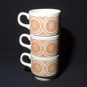 Könnte beinhalten: Drei weiße Keramik-Kaffeetassen, die übereinander gestapelt sind. Jede Tasse hat ein braunes und weißes geometrisches Muster aus konzentrischen Kreisen.