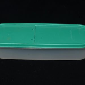 Pode incluir: Um recipiente retangular para armazenamento de alimentos com uma base translúcida e uma tampa verde-azulado. A tampa tem uma área rebaixada e uma aba para facilitar a abertura. O recipiente foi projetado para armazenar alimentos.