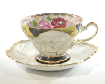 May include: A white porcelain teacup and saucer with a floral design. The cup has a black rim and gold accents. The saucer has a leaf design with gold accents.