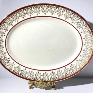 May include: Oval serving platter with a cream-colored center and a decorative border. The border features a red band with gold and cream-colored ornate designs. The platter is displayed on a decorative gold-colored stand.