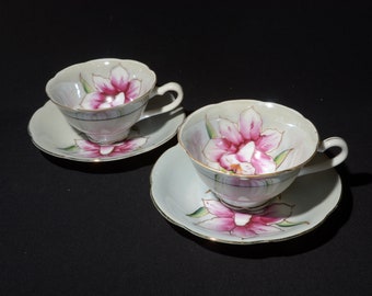 以下が含まれることがあります： ピンクの花柄と金色のアクセントが施された白いティーカップが2つ。カップは同じ花柄のソーサーに乗っています。