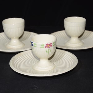 May include: Set of three white ceramic egg cups with floral designs. Each cup has a matching saucer with a fluted edge.