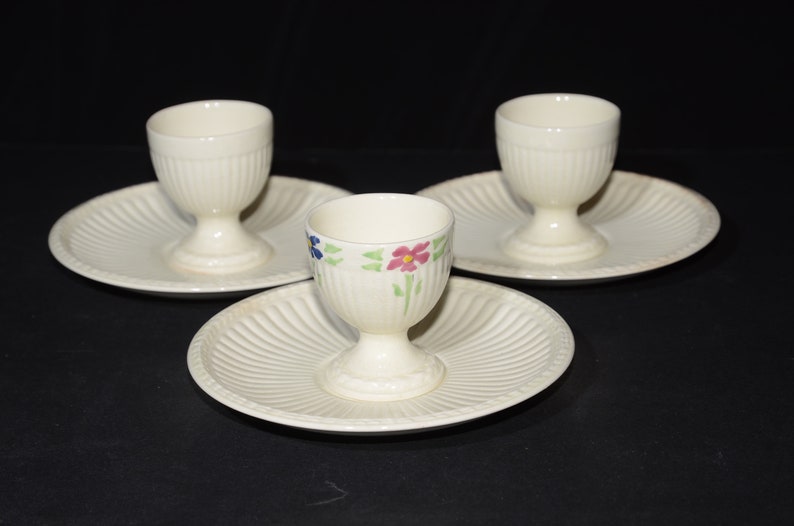 May include: Set of three white ceramic egg cups with floral designs. Each cup has a matching saucer with a fluted edge.