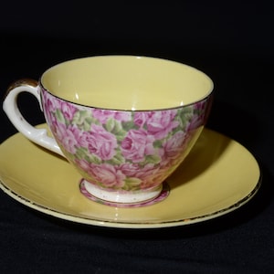 May include: A yellow teacup and saucer with a floral pattern of pink roses and green leaves. The cup and saucer have a gold trim.