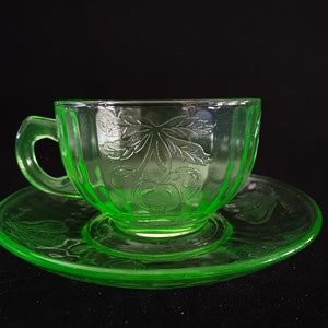 Può includere: Un set da tè in vetro verde con un design a ciliegie. La tazza e il piattino sono entrambi traslucidi e hanno un aspetto delicato e vintage.