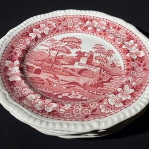 Może przedstawiać: Stos białych i czerwonych ceramicznych talerzy. Talerze mają czerwony kwiatowy i krajobrazowy wzór z mostem i sceną zamkową. Obręcze mają falisty brzeg z białą obwódką.