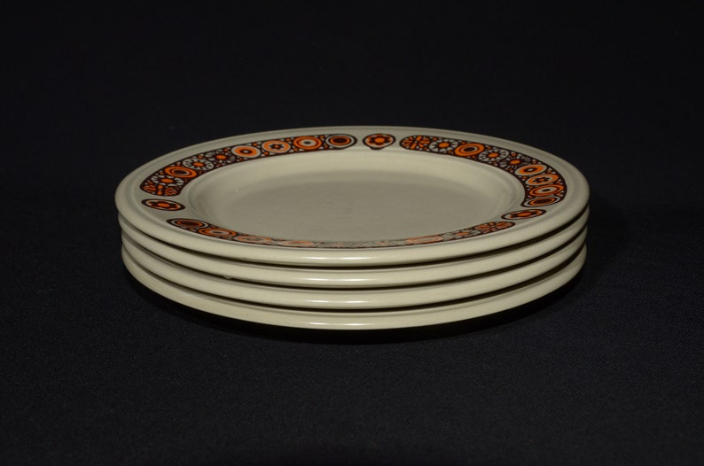 May include: A stack of four white ceramic plates with a brown and orange geometric pattern around the rim.