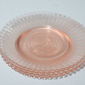 Puede incluir: Una pila de cuatro platos de vidrio rosa. Cada plato tiene un borde festoneado y un dise&ntilde;o texturizado y acanalado que irradia desde el centro. Los platos son de un color rosa suave y transl&uacute;cido.
