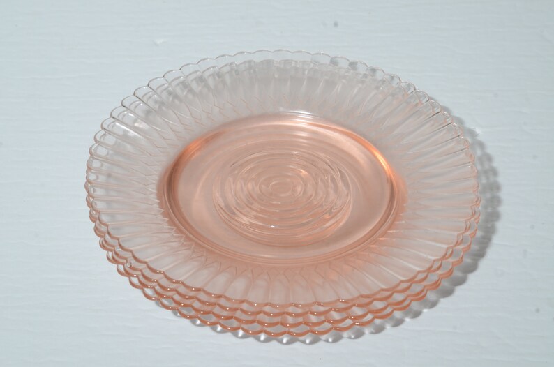 Puede incluir: Una pila de cuatro platos de vidrio rosa. Cada plato tiene un borde festoneado y un dise&ntilde;o texturizado y acanalado que irradia desde el centro. Los platos son de un color rosa suave y transl&uacute;cido.