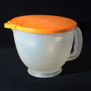 Könnte beinhalten: Eine transparente Kunststoff-Rührschüssel mit einem orangefarbenen Deckel. Die Schüssel hat einen Griff und Messmarkierungen in Millilitern. Der Deckel hat eine Lasche zum einfachen Öffnen. Ein Küchenutensil zum Messen und Mischen.