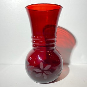 May include: A small, red glass vase with a rounded base and a flared top. The vase has a clear, etched design of a flower on the front.