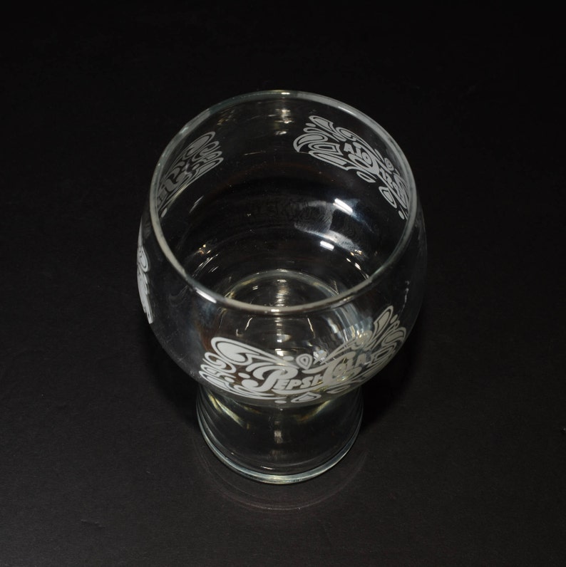 May include: A clear glass drinking glass with an etched design of the Pepsi logo and waves. The glass has a wide base and a narrow top.