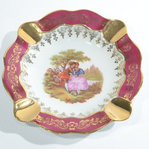 May include: A square porcelain ashtray with a gold rim and a pink and gold floral design. The center of the ashtray features a painted scene of a couple in a garden.
