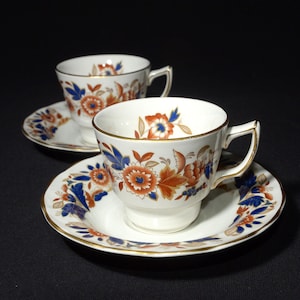 Puede incluir: Dos tazas de té blancas con motivos florales azules y naranjas y ribetes dorados. Las tazas de té están sobre platillos a juego con el mismo motivo floral.