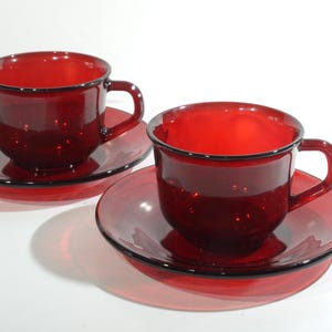 Könnte beinhalten: Zwei passende rote Teetassen und Untertassen aus Glas. Die Tassen haben eine abgerundete Form mit Henkel und stehen auf passenden Untertassen. Jedes Teil hat einen dunklen Rand. Das Set wird auf einer weißen Oberfläche präsentiert.
