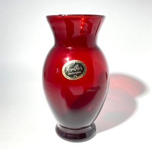 May include: A translucent ruby red glass vase with a flared rim and a rounded body. An oval sticker on the front features the text Royal Ruby Anchorglass Lancaster Ohio U.S.A. in gold lettering on a black background, indicating the brand.