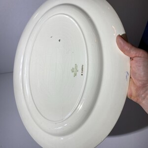 May include: An oval, off-white ceramic serving platter. The underside features a raised rim and a central, slightly recessed area. There is a small, faded emblem and text near the rim. The platter is held by a hand.
