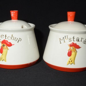 以下が含まれることがあります： 赤い蓋と雄鶏のイラストが描かれた白いセラミック製の調味料入れが2つ。左側の容器には「ケチャップ」と書いてあり、右側の容器には「マスタード」と書いてあります。