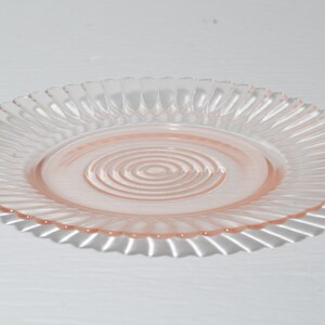 Puede incluir: Un plato de cristal transparente y rosa con un borde acanalado y un dise&ntilde;o de c&iacute;rculos conc&eacute;ntricos en el centro. El plato es redondo y proyecta una sombra sobre la superficie blanca de abajo. Este plato vintage es perfecto para servir aperitivos o postres.