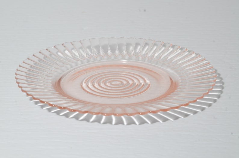 Puede incluir: Un plato de cristal transparente y rosa con un borde acanalado y un dise&ntilde;o de c&iacute;rculos conc&eacute;ntricos en el centro. El plato es redondo y proyecta una sombra sobre la superficie blanca de abajo. Este plato vintage es perfecto para servir aperitivos o postres.
