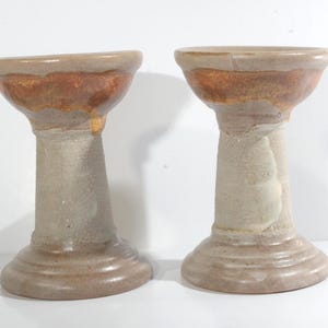 May include: Two ceramic candle holders with a light brown glaze and a darker brown rim. The holders have a wide base and a narrow neck.