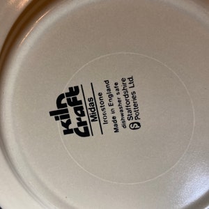 May include: Close-up of the bottom of a beige ceramic plate with black text that reads "Kiln Craft Midas Ironstone Made in England Dishwasher safe Staffordshire Potteries Ltd."