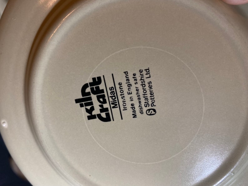 May include: Close-up of the bottom of a beige ceramic plate with black text that reads "Kiln Craft Midas Ironstone Made in England Dishwasher safe Staffordshire Potteries Ltd."