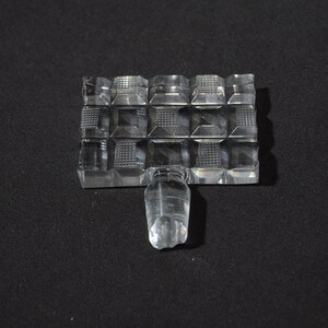 May include: A clear glass bottle stopper with a square base and a textured design. The stopper has a round top and a small, clear glass knob.