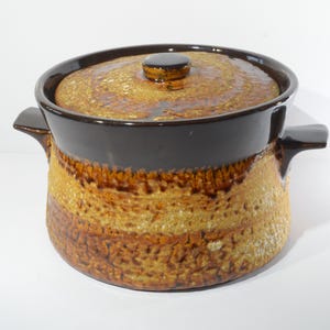 Puede incluir: Una fuente de cerámica con tapa. La fuente tiene un borde y asas de color marrón oscuro, con un diseño texturizado y en capas en tonos marrones y beige. La tapa tiene un pequeño pomo.