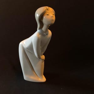May include: A white porcelain figurine of a girl with blonde hair, wearing a white dress. She is looking up and to the right, with a slight smile on her face.