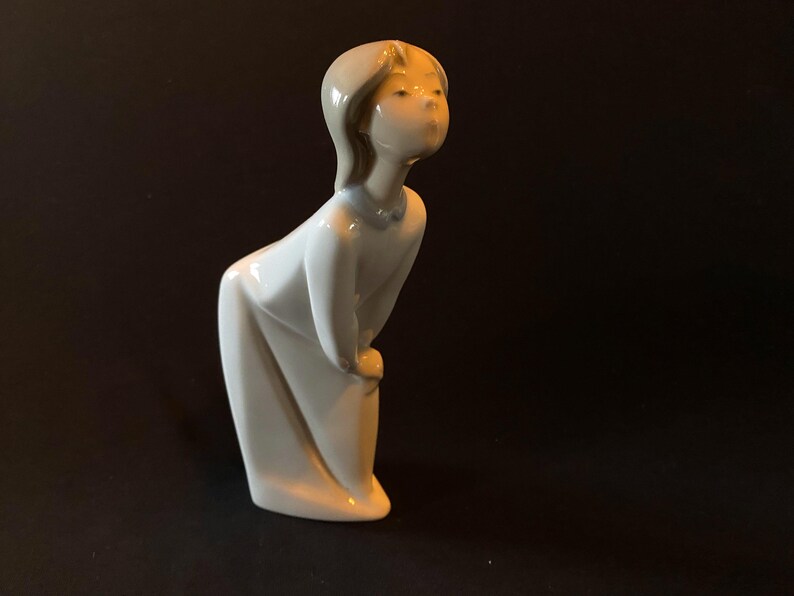 May include: A white porcelain figurine of a girl with blonde hair, wearing a white dress. She is looking up and to the right, with a slight smile on her face.