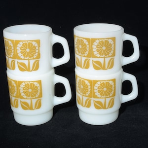May include: Four white ceramic mugs with a yellow floral design. Each mug features a square pattern with a flower and leaves. The mugs are stacked in pairs. The mugs are suitable for hot drinks.