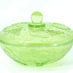 Peut inclure: Un bonbonnière en verre vert avec un couvercle. Le plat est rond avec un motif décoratif de raisins et de feuilles. Le couvercle a un bouton rond. Le plat est en verre vert translucide.