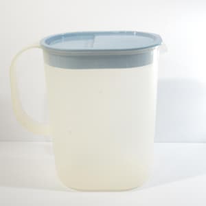Può includere: Una caraffa di plastica trasparente con un coperchio blu chiaro. La caraffa ha un manico e un beccuccio.