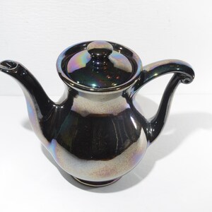 May include: A small, iridescent black teapot with a lid. The teapot has a rounded body and a curved spout. The handle is also curved and has a decorative finial.