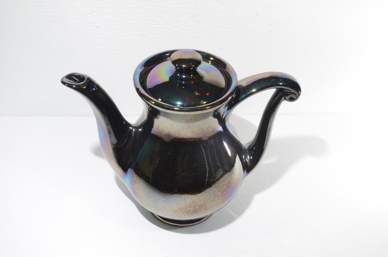 May include: A small, iridescent black teapot with a lid. The teapot has a rounded body and a curved spout. The handle is also curved and has a decorative finial.