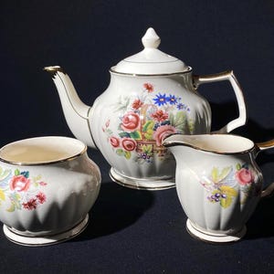 Puede incluir: Una tetera, una jarra de crema y un azucarero blancos con un patrón floral. La tetera tiene un borde dorado y una tapa. La jarra de crema y el azucarero también tienen un borde dorado.