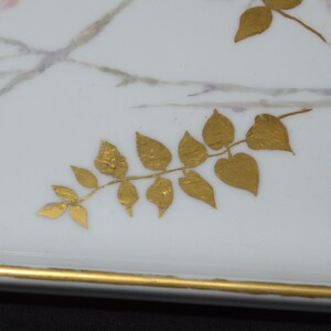 May include: A rectangular white ceramic tray with a gold rim and gold leaf accents. The tray features delicate pink and purple floral designs and gold painted leaves, creating an elegant aesthetic.