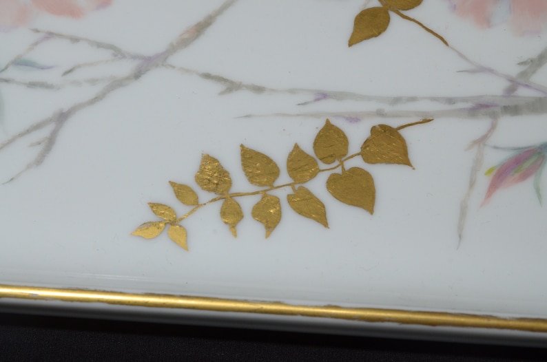 May include: A rectangular white ceramic tray with a gold rim and gold leaf accents. The tray features delicate pink and purple floral designs and gold painted leaves, creating an elegant aesthetic.