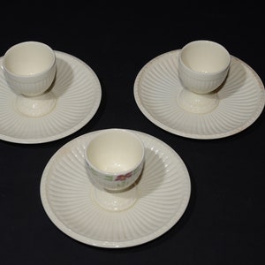 May include: Three white ceramic egg cups with matching plates. The egg cups have a floral design and are on a black background.