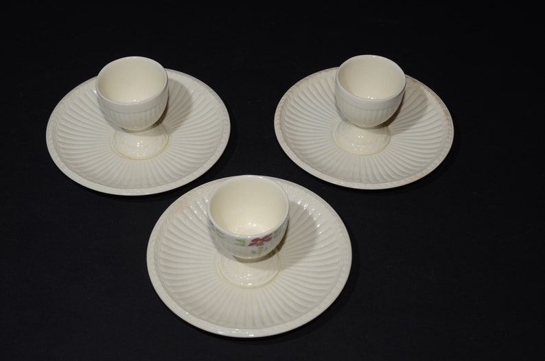 May include: Three white ceramic egg cups with matching plates. The egg cups have a floral design and are on a black background.