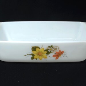 May include: A rectangular white milk glass baking dish with rounded corners and integrated side handles. The exterior front features a floral decal with a large yellow flower, a smaller orange flower, and green leaves against the white surface.