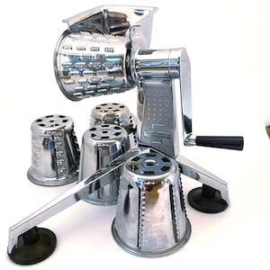 May include: A chrome-plated rotary food grater with a hand crank and black handle. The device features a tripod base with black suction feet and includes three interchangeable cylindrical stainless steel grating drums with various cutting patterns.