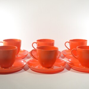 May include: Set of six orange ceramic teacups and saucers. The cups and saucers are all the same size and shape. They are arranged in a row on a white surface.