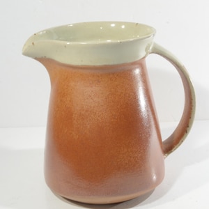 May include: A brown ceramic pitcher with a light beige rim and handle. The pitcher has a slightly flared shape and a smooth, glazed finish.
