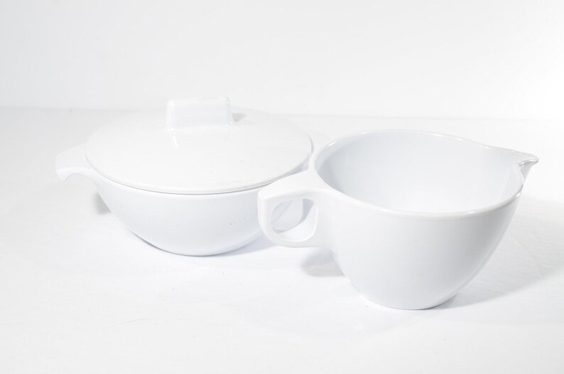 May include: White ceramic sugar bowl and creamer set. The sugar bowl has a lid with a square handle. The creamer has a handle and a spout.