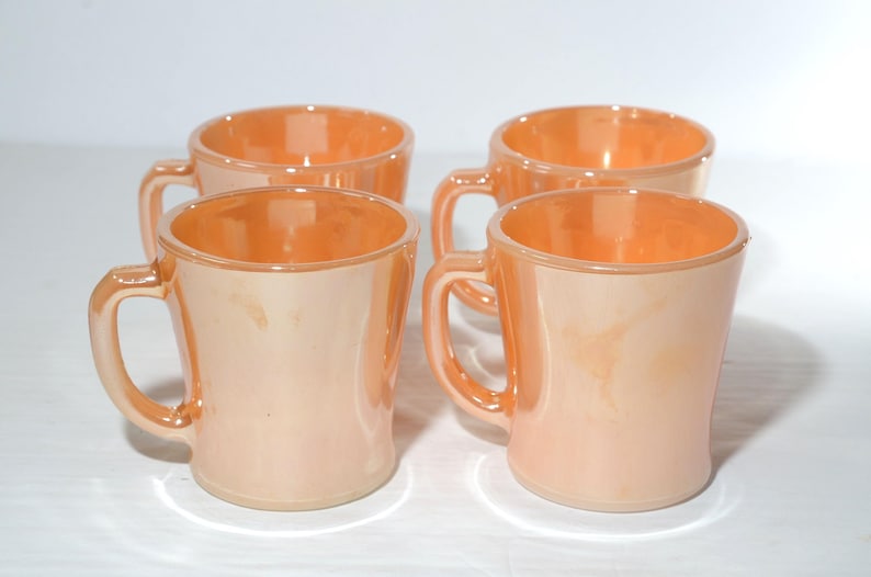 May include: Four peach colored ceramic mugs with white interiors and handles. The mugs are arranged in a square shape on a white surface.